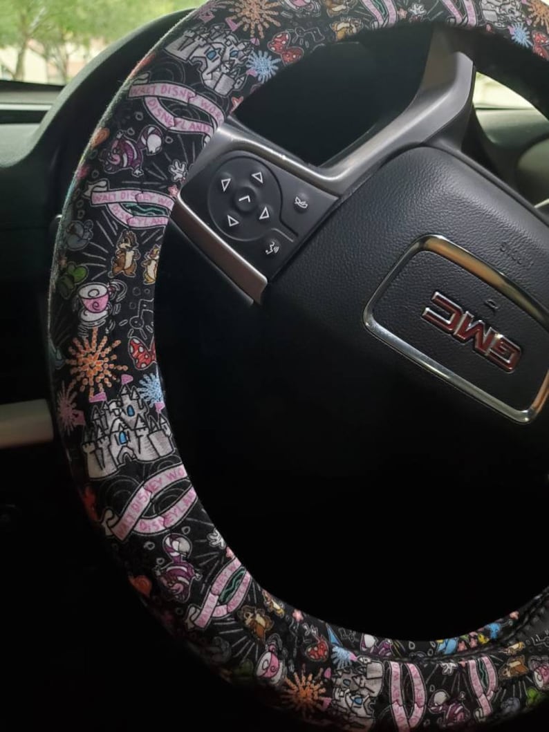 Puede incluir: Funda de volante negra con un estampado de Disney colorido que presenta personajes como Mickey Mouse, Minnie Mouse y el castillo de Cenicienta. El texto "Walt Disney World" est&aacute; impreso en la funda.