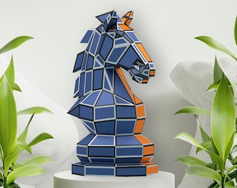 Puede incluir: Una escultura geométrica de un caballo de ajedrez azul y naranja. La estatua se exhibe sobre un pedestal blanco, rodeada de plantas verdes. El caballo presenta un patrón de mosaico con detalles en azul oscuro y naranja.