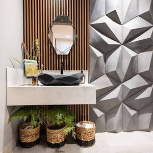 May include: Modern bathroom design featuring a black geometric sink, a black framed mirror, and a textured gray wall. The sink sits on a white countertop with plants in woven baskets below. The background has vertical wood paneling.