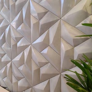 May include: A wall with a geometric design composed of repeating, three-dimensional, triangular shapes. The tiles are a light, neutral color, creating a textured surface. Green plant leaves are visible in the foreground.