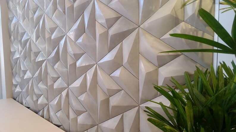 May include: A wall with a geometric design composed of repeating, three-dimensional, triangular shapes. The tiles are a light, neutral color, creating a textured surface. Green plant leaves are visible in the foreground.