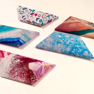 May include: Five triangular gift boxes with various designs. One box has a blue and white winter snowflake pattern. Another features a watercolor ocean scene. The others have abstract floral and holiday patterns. The boxes are arranged on a white surface.