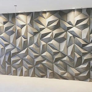 May include: A large wall panel with a geometric, three-dimensional design. The panel is composed of numerous triangular and polygonal shapes in varying shades of gray, creating a textured surface. The wall is illuminated by recessed lighting.