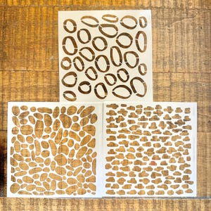 May include: Three square white plastic stencils featuring distinct patterns. The top stencil displays large open oval shapes. The bottom left stencil shows a dense arrangement of irregular pebble-like forms. The bottom right stencil features small cloud-like shapes.