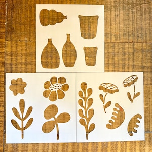 May include: Three white sheets featuring brown cut-out shapes. The top sheet displays vases and pots. The bottom left sheet shows a flower, a leaf sprig, and a clover-like plant. The bottom right sheet depicts a tall leaf, two flowers, and two abstract shapes.