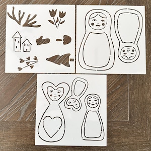 May include: Three white stencils with brown cut-out designs. One stencil features various nature-themed shapes, including a tree, houses, and flowers. The other two stencils feature Matryoshka doll designs, with one doll having a heart shape.