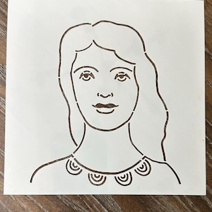 May include: A white stencil featuring a woman's face with long hair and a necklace. The design is cut out, creating a negative space image. The stencil is on a wooden surface.