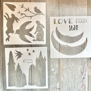 May include: A selection of white stencils with diverse designs. One stencil displays the text "LOVE MOON STAR" above two crescent moon shapes. Other stencils feature a bird on a branch, a rabbit, a flying bird, stars, and a castle.