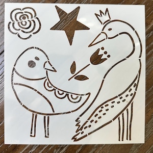 May include: A square white stencil featuring cutouts of a round bird, a swan wearing a crown, a flower, a five-pointed star, and a rose. The design elements are arranged across the surface, creating a template for crafting projects.