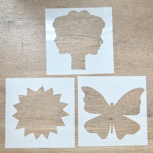 May include: Three white plastic stencils on a wooden surface. The stencils feature a profile of a person, a starburst shape, and a butterfly. The stencils are square and have cut-out designs.