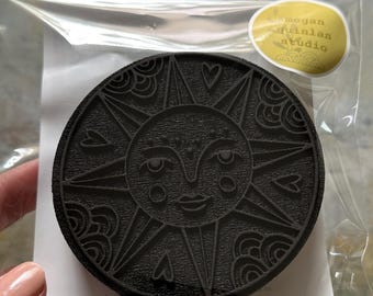 May include: A circular black stamp featuring an embossed sun face design with rays, clouds, and small hearts. The stamp is enclosed in a clear plastic bag with a yellow sticker in the corner that reads megan quinlan studio.