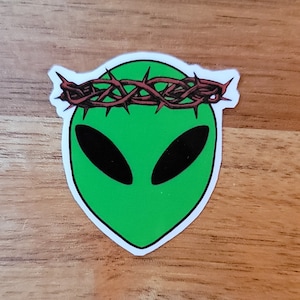 May include: A sticker featuring a green alien head with large black oval eyes. The alien wears a crown of thorns in shades of red and brown. The sticker is on a wooden surface.