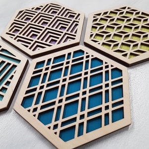 May include: Four hexagonal coasters with geometric patterns cut out of the wood. The coasters are made of light wood with a dark brown edge and have different coloured backgrounds: teal, blue, purple, and yellow.
