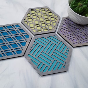 Puede incluir: Juego de cuatro posavasos hexagonales de madera con diseños geométricos. Cada posavasos presenta un color diferente: azul, turquesa, amarillo y morado. Los posavasos tienen un borde gris y están dispuestos sobre una superficie blanca.