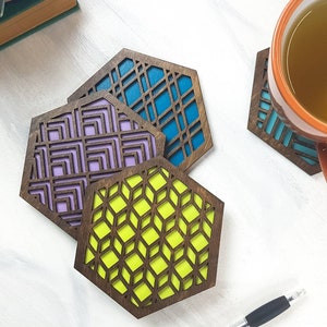 May include: Three wooden coasters with geometric designs. The coasters are in shades of purple, blue, and green. The coasters are hexagonal in shape.