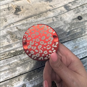 Puede incluir: Un objeto circular rojo con un diseño de estampado de leopardo en gris claro. El objeto está sostenido por una mano con esmalte de uñas rosa. El fondo es una superficie de madera.