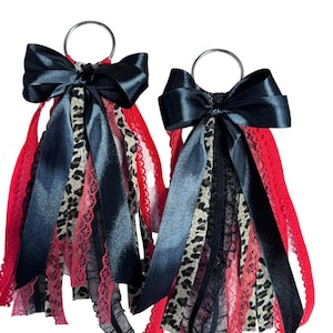 Op de afbeelding: Twee decoratieve sleutelhangers met zwarte satijnen strikken en bungelende linten. De linten bevatten rood kant, zwart kant en stof met luipaardprint. Elke sleutelhanger heeft een zilveren metalen ring.