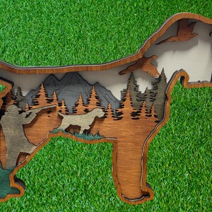 May include: A wooden wall art piece shaped like a dog, with a hunter and a dog in a forest scene with mountains in the background. The dog is brown and the hunter is wearing a brown jacket and pants. The forest is green and the mountains are gray.