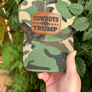 May include: Camouflage baseball cap with a brown leather patch that reads "COWBOYS FOR TRUMP" in black lettering.