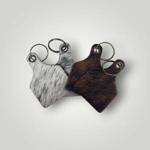 May include: Four tag-shaped keychains made of textured animal hide. Two keychains feature a white and black speckled pattern, while the other two display a solid dark brown color. Each piece includes a circular metal ring attached at the top.