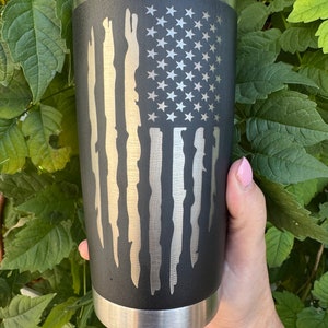 May include: A black and silver stainless steel tumbler with a distressed American flag design. The flag is in shades of gray and white.