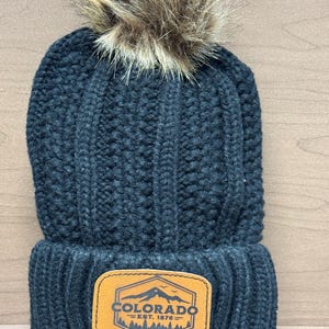 Pode incluir: Boina de malha preta com um pompom de pele sintética marrom e um remendo de couro marrom que diz "Colorado - Est. 1876 -" com um gráfico de montanha e árvores.