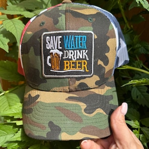 Puede incluir: Gorra de béisbol de camuflaje con una parte trasera de malla y un parche que dice "SAVE WATER DRINK BEER" en blanco, azul y amarillo. La gorra tiene una visera curva y un botón marrón en la parte superior.