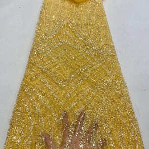 May include: A close-up of a yellow beaded fabric with a geometric pattern. The fabric is covered in small, shiny beads, creating a textured surface. The design features vertical and diagonal lines, and a small piece of matching fabric is at the top.