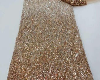 May include: A shimmering, beaded fabric in shades of gold and champagne. The fabric is densely covered with sequins, creating a textured, sparkling effect. The material is folded, revealing a glimpse of a sheer, matching underlay.
