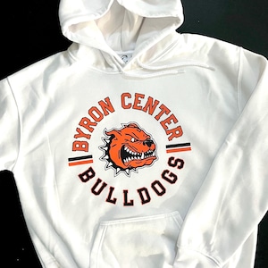May include: White hooded sweatshirt with a circular graphic of a snarling orange bulldog with black outlines. The graphic reads "BYRON CENTER BULLDOGS" in black and orange text.