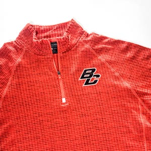 May include: A red textured quarter-zip pullover with the letters "BC" in black and white on the right chest. The shirt has a high collar and a zipper that extends down the chest. The fabric has a subtle pattern.