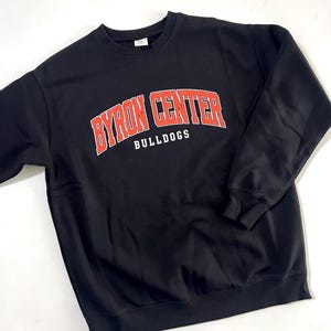 May include: Black crewneck sweatshirt with the text "BYRON CENTER" in red arched lettering and the word "BULLDOGS" in white below. The sweatshirt is a solid black color.