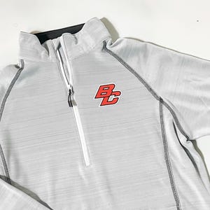 May include: A light gray quarter-zip athletic top with a black collar and a red and black logo on the chest. The top has a white zipper and black stitching along the seams.