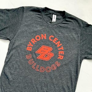 May include: A heather gray t-shirt with a red circle graphic. The graphic says "Byron Center Bulldogs" with the letters "BC" in a larger font inside the circle.