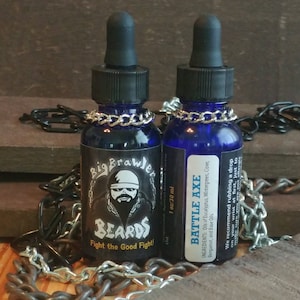 May include: Two 1 ounce bottles of beard oil. The bottles are blue with black screw-on caps and silver chains around the necks. The labels are black with white text. The left bottle says "Big Brawler Beards" and "Fight the Good Fight!" The right bottle says "Battle Axe" and lists the ingredients.