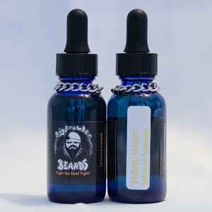 May include: Two blue glass bottles with droppers, each containing beard oil. The bottle on the left has a label that says "Big Brawler Beards" and "Fight the Good Fight!" The bottle on the right has a label that says "Tropic Smoke Tobacco Banana".