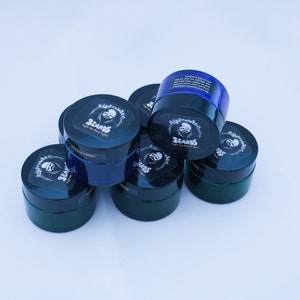 May include: A group of six small, round, blue and green jars with black lids. Each jar has a label with the words "Big Brawlers Beards" and a cartoon image of a bearded man.
