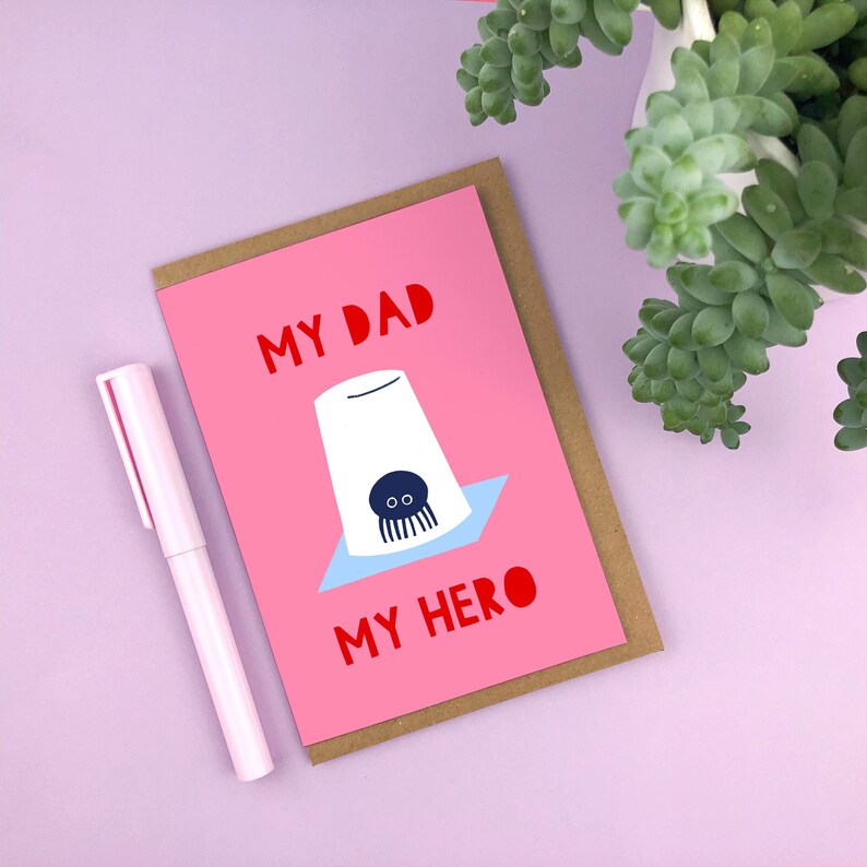 Puede incluir: Una tarjeta rosa con el texto "My Dad My Hero" en letras rojas. Una ilustraci&oacute;n de dibujos animados de una ara&ntilde;a en una taza est&aacute; en la tarjeta.