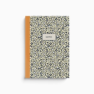 Puede incluir: Un cuaderno beige con una cubierta de estampado de leopardo negro y blanco y una encuadernación naranja. La portada tiene la palabra "NOTES" impresa en un rectángulo negro.