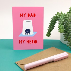Puede incluir: Tarjeta rosa con el texto "My Dad My Hero" en letras rojas. Una ilustraci&oacute;n de dibujos animados de un pulpo en una taza blanca est&aacute; en el centro de la tarjeta.