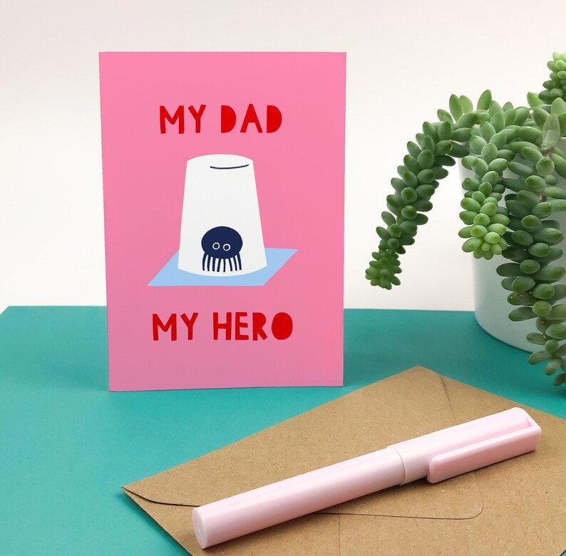 Puede incluir: Tarjeta rosa con el texto "My Dad My Hero" en letras rojas. Una ilustraci&oacute;n de dibujos animados de un pulpo en una taza blanca est&aacute; en el centro de la tarjeta.