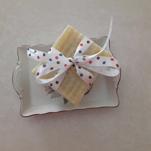 May include: A bar of soap wrapped in a white ribbon with red, blue and green polka dots. The soap is sitting on a white porcelain dish with a floral design.
