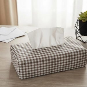 Peut inclure: Une housse de boîte à mouchoirs rectangulaire avec un motif à carreaux vichy marron et blanc. La housse est en tissu et recouvre une boîte à mouchoirs standard, avec un mouchoir blanc dépassant par l'ouverture supérieure du contenant.