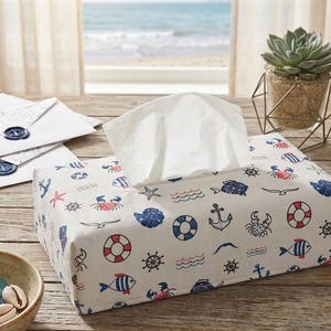 Peut inclure: Une housse de boîte à mouchoirs rectangulaire en tissu blanc avec un motif nautique répétitif en rouge, bleu et noir. L'imprimé comprend des illustrations de crabes, poissons, ancres, bouées, roues de gouvernail, étoiles de mer et vagues.