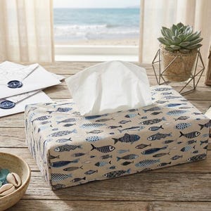 Peut inclure: Un distributeur de mouchoirs rectangulaire avec un fond beige et un motif de poissons bleus et marine. Des mouchoirs blancs dépassent du dessus. La scène comprend une plante succulente, une photo de phare encadrée et un bol décoratif sur une table en bois.