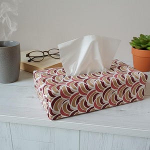 Peut inclure: Une boîte à mouchoirs rectangulaire recouverte d'un tissu à motif répétitif de demi-cercles superposés dans des tons marron, beige et blanc. La housse contient une pile de mouchoirs en papier blancs qui dépassent par l'ouverture supérieure.