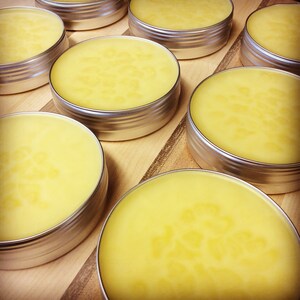 May include: Several small round containers of yellow balm, each with a silver lid, are arranged on a wooden surface.