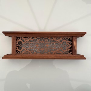 Puede incluir: Una estantería rectangular de madera con un diseño decorativo tallado. La estantería es de color marrón cálido y presenta un intrincado patrón calado en el centro. La estantería está montada en una pared blanca.