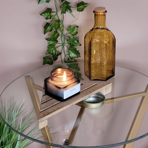 Könnte beinhalten: Ein Glas-Kerzengefäß mit einem dreieckigen Holzständer, einer brennenden Kerze darin und einem Etikett mit der Aufschrift "Jasmine & Fig" auf einem Glastisch mit einer dekorativen, bernsteinfarbenen Glasflasche.