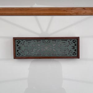 Puede incluir: Una rejilla de ventilación rectangular con un marco de madera marrón oscuro que rodea un inserto metálico de color verde salvia. El inserto presenta un patrón simétrico y complejo de volutas cortadas con láser, con líneas fluidas.